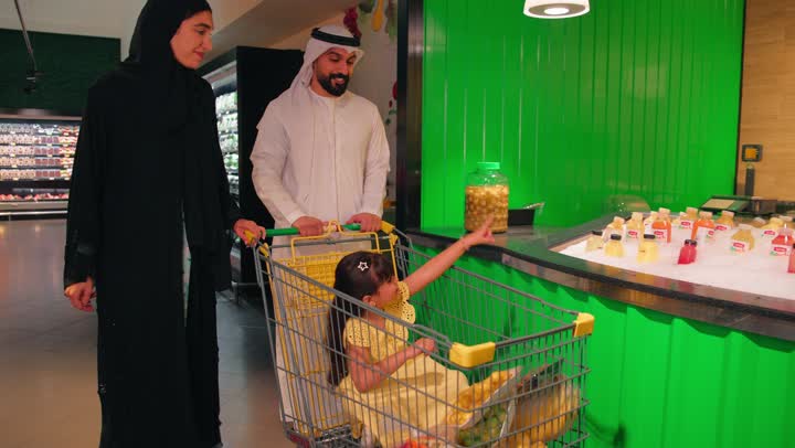 An Arab Gulf Emirati family dressed in traditional attire inside the supermarket is selecting bottles of fresh juice from the beverage section, pointing to something, the concept of family shopping and buying daily necessities, concern for healthy nutrition for children, gestures of happiness and joy, spending enjoyable time with the family while shopping.
