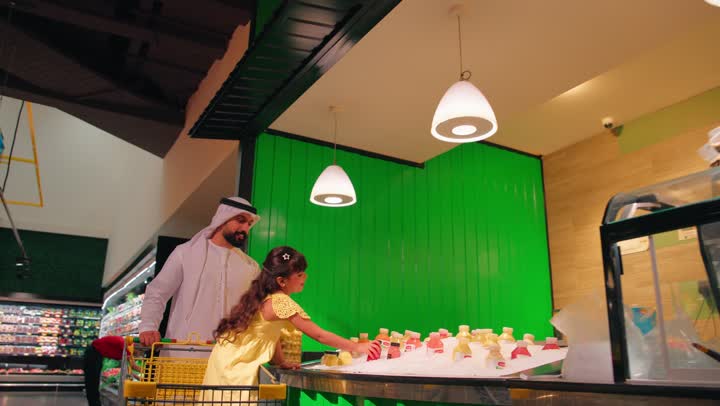 Spending a good time inside the mall, an Emirati Gulf Arab man wearing traditional attire with his daughter in the juice section of the supermarket, choosing fresh drinks and juices from the store, the concept of family shopping and buying daily necessities, focusing on healthy nutrition and a healthy lifestyle.