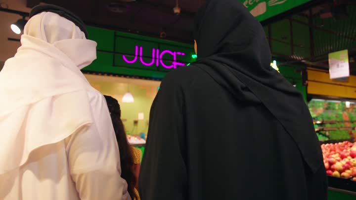 A rear view of an Emirati Gulf Arab family dressed in traditional attire heading to buy fresh juices inside the supermarket, family shopping and choosing healthy drinks from the juice section, having a good time inside the store.