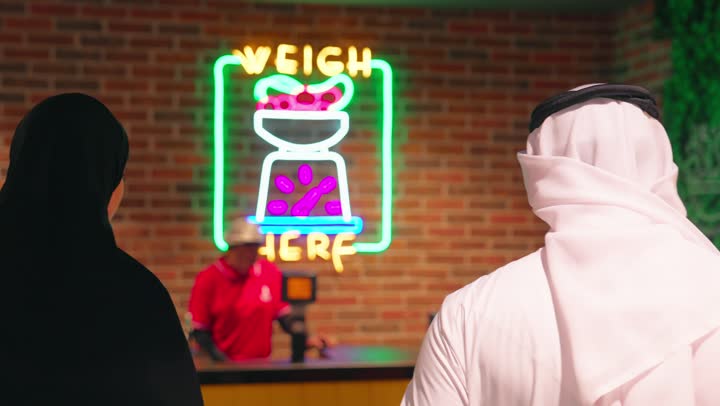 التسوق داخل السوبر ماركت، تصوير من الخلف لعائلة عربية خليجية اماراتية ترتدي الزي التقليدي تقوم بوزن الفواكه والخضروات الطازجة في قسم المنتجات، موظفة المتجر تساعد العملاء، مفهوم شراء الاحتياجات اليومية، التسوق العائلي والاستمتاع بوقت داخل المتجر