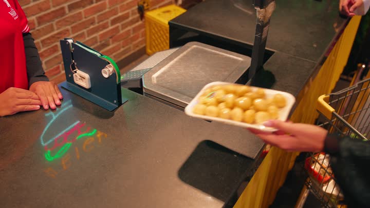 Buying fresh fruits inside the supermarket, a close-up shot of the hand of an Emirati Gulf Arab woman holding a pack of red grapes and a pack of fresh fruit as she pays for her purchases at the cashier, the accounting employee is registering the products on the electronic scale, the concept of shopping and buying daily necessities.