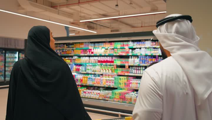 An Arab Gulf Emirati family is shopping and buying household supplies from the supermarket. An Arab Gulf Emirati man wearing a kandura and ghutrah is pushing the shopping cart accompanied by his wife and daughter. The concept of family cooperation and family bonding while shopping, purchasing daily necessities inside the store.