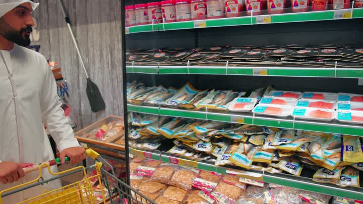التسوق المنزلي وتوفير الاحتياجات اليومية، تصوير مقربلرجل عربي خليجي اماراتي يرتدي الزي التقليدي يمسك بيدية عربة التسوق  يختار عبوة مأكولات متبلة من ثلاجة قسم الأسماك في السوبر ماركت