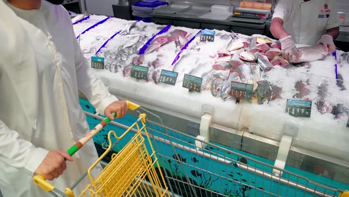 Buying fresh seafood products, preparing and selling fresh fish in the seafood section of the supermarket, an Emirati Gulf Arab man wearing traditional attire is pulling a shopping cart and selecting the appropriate type of fish, the concept of customer service inside the store.