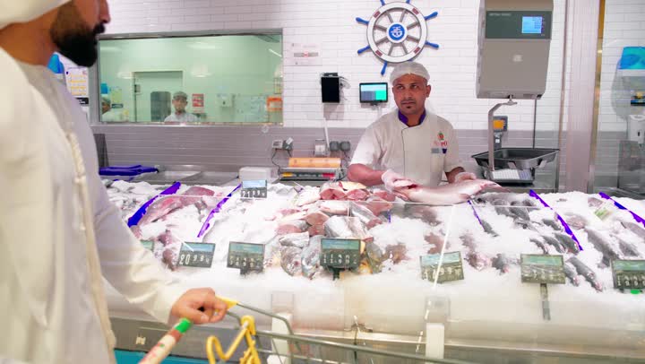 تجهيز وبيع الأسماك الطازجة داخل قسم المأكولات البحرية في السوبر ماركت، رجل عربي خليجي اماراتي يرتدي الزي التقليدي يختار نوع السمك المناسب، شراء المنتجات البحرية الطازجة، مفهوم خدمة العملاء داخل المتجر