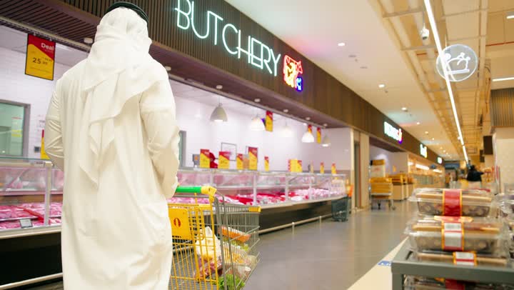 اختيار اللحوم الطازجة من قسم الجزارة داخل السوبر ماركت، رجل عربي خليجي اماراتي يرتدي الثوب التقليدي يقف أمام ثلاجة عرض اللحوم ويدفع عربة التسوق أثناء طلب احتياجاته من الموظف، عامل يقوم بتجهيز الطلب وتقديمه للعميل، مفهوم شراء المواد الغذائية اليومية والتسوق المنزلي، خدمة العملاء داخل متجر