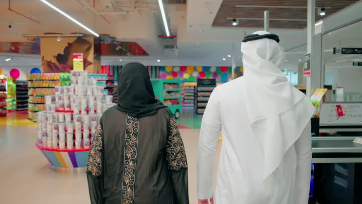 جولة تسوق داخل السوبر ماركت ، تصوير من الخلف لزوجان عربيان خليجيان اماراتيان يسيران  بين الممرات بعد الانتهاء من اختيار المنتجات، مفهوم التسوق العائلي والتخطيط لشراء الاحتياجات اليومية، الاستعداد للدفع ومتابعة قائمة المشتريات