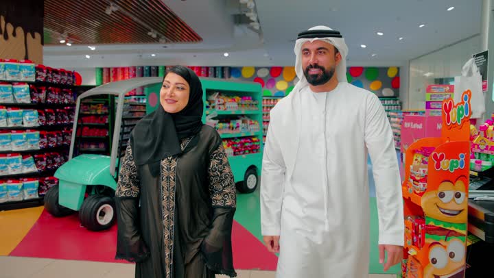 Walking inside the supermarket during family shopping, an Emirati Gulf Arab woman in a hijab is pointing with her hand towards the products inside the store accompanied by her husband, planning to buy daily necessities and identifying the required products, the concept of conscious shopping and choosing consumer goods.