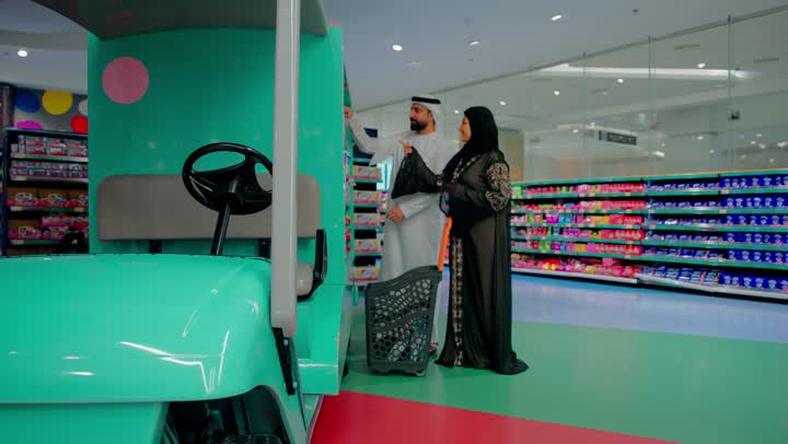 مفهوم شراء احتياجات الأطفال والاستعداد للمناسبات والهدايا، التسوق العائلي داخل قسم ألعاب الأطفال في السوبر ماركت، زوجان عربيان خليجيان اماراتيان يرتديان الزي التقليدي يقومان باختيار ألعاب ودمى للأطفال، المشاركة في اختيار المنتجات، مقارنة الألعاب ومعرفة الأسعار داخل المتجر