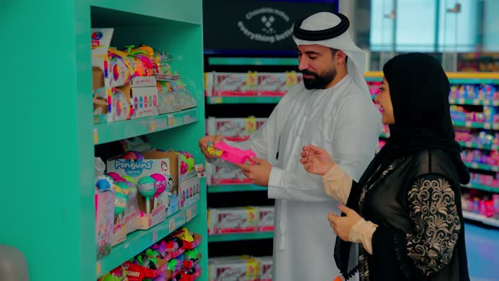 التسوق العائلي داخل قسم ألعاب الأطفال في السوبر ماركت، زوجان عربيان خليجيان اماراتيان يرتديان الزي التقليدي يقومان باختيار ألعاب ودمى للأطفال، المشاركة في اختيار المنتجات، مقارنة الألعاب ومعرفة الأسعار داخل المتجر، مفهوم شراء احتياجات الأطفال والاستعداد للمناسبات والهدايا
