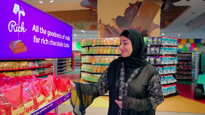 اختيار حلويات الأطفال وشراء المستلزمات العائلية داخل السوبر ماركت، امرأة عربية خليجية اماراتية ترتدي العباءة والحجاب تقوم بإعطاء زوجها كيس من الحلويات، التعاون بين الزوجين وتبادل المنتجات أثناء التسوق، مفهوم التسوق العائلي واختيار المنتجات المناسبة قبل الشراء