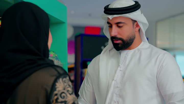 اختيار حلويات الأطفال داخل السوبر ماركت،تصوير مقرب لرجل عربي خليجي اماراتي  يرتدي الثوب التقليدي يقف برفقة زوجتة يقوم بإمساك حلوى للأطفال ، شراء المستلزمات والمنتجات الخاصة بالأطفال، مفهوم التسوق العائلي واختيار المنتجات بعناية قبل الشراء