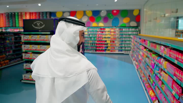 تفقد المنتجات الغذائية داخل السوبر ماركت واختيار الوجبات الخفيفة، رجل عربي خليجي اماراتي يرتدي الثوب التقليدي يقوم بإمساك عبوة سناكس يقرأ تفاصيل المنتج قبل الشراء،  التسوق الواعي ومقارنة السلع الغذائية داخل المتجر، مفهوم شراء الاحتياجات اليومية والاستهلاك