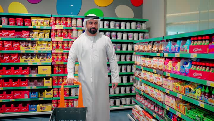 اختيار منتجات الشوكولاتة والحلويات داخل السوبر ماركت، مفهوم شراء الاحتياجات اليومية والاستهلاك في الامارات العربية، رجل عربي خليجي اماراتي يرتدي الثوب التقليدي يقوم بإمساك عبوة شوكولاتة و يضعها فى سلة التسوق،  التسوق الواعي ومقارنة السلع الغذائية داخل المتجر