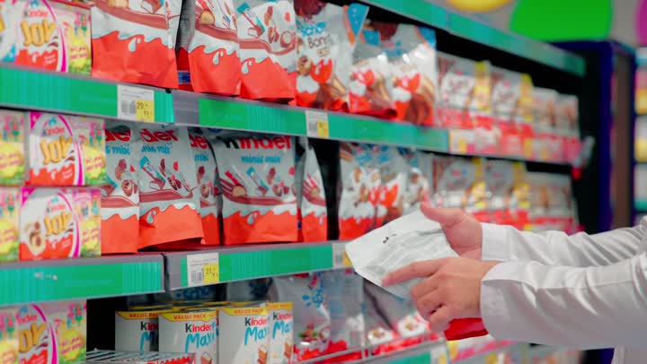 مفهوم شراء الاحتياجات اليومية والاستهلاك في الامارات العربية، اختيار منتجات الشوكولاتة والحلويات داخل السوبر ماركت، تصوير مقرب ليد رجل عربي خليجي اماراتي يرتدي الثوب التقليدي يقوم بإمساك عبوة شوكولاتة،  التسوق الواعي ومقارنة السلع الغذائية داخل المتجر