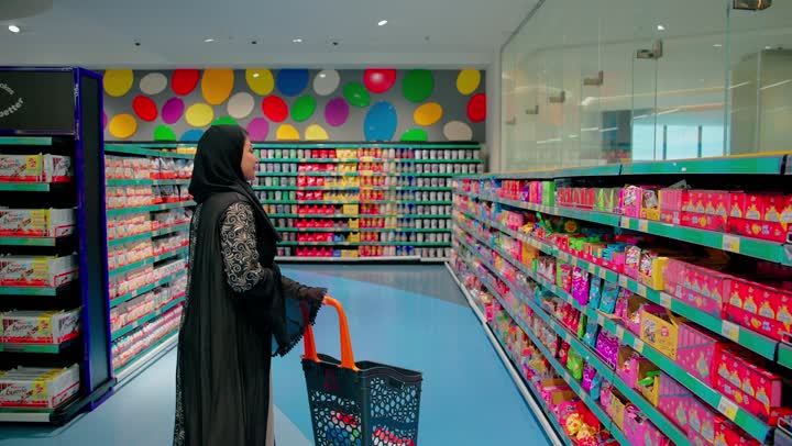 Using a shopping cart and buying daily necessities, shopping inside the supermarket and carefully selecting food products, an Arab Gulf Emirati woman wearing an abaya and hijab stands in the snack and sweets aisle holding a chocolate package and reading the product details before purchasing.