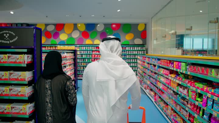 مفهوم شراء الاحتياجات اليومية واختيار المنتجات بعناية، التسوق العائلي داخل السوبر ماركت ، تصوير من الخلف لرجل عربي خليجي اماراتي يرتدي الثوب التقليدي برفقة زوجته يتجولان بين ممرات المتجر ويدفعان عربة التسوق أثناء تفقد رفوف المنتجات الغذائية