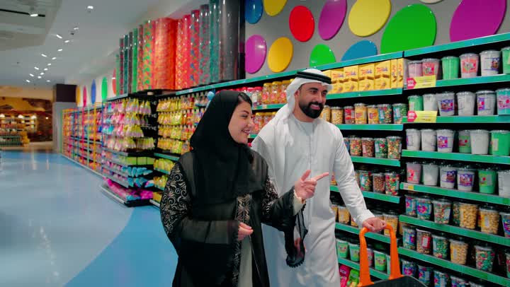 The concept of carefully selecting products and daily consumption, a tour inside the supermarket to buy daily necessities, an Emirati Gulf Arab man wearing traditional attire holding a shopping cart alongside his wife, talking and checking the shelves of food products and snacks, family shopping and comparing goods before purchase.
