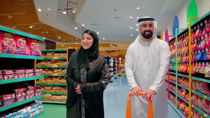 جولة داخل السوبر ماركت لشراء الاحتياجات اليومية، رجل عربي خليجي اماراتي يرتدي الثوب التقليدي يمسك عربة التسوق برفقة زوجته يتحدثان ويقومان بتفقد رفوف المنتجات الغذائية والوجبات الخفيفة، التسوق العائلي ومقارنة السلع قبل الشراء، مفهوم اختيار المنتجات بعناية والاستهلاك اليومي