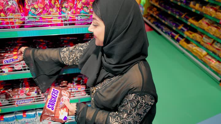 مفهوم التسوق داخل المتجر وشراء الاحتياجات اليومية، تصوير مقرب لامرأة عربية خليجية اماراتية ترتدي العباءة السوداء والحجاب تقوم باختيار وشراء الشوكولاتة والحلويات من رفوف السوبر ماركت، السناكات والمنتجات الغذائية المتنوعة