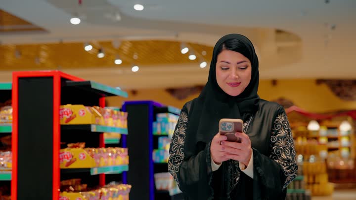Gestures of happiness and joy, a close-up shot of an Arab Gulf Emirati woman wearing a black abaya and hijab standing inside a supermarket using a mobile phone, browsing social media and communicating with family and friends via mobile, the concept of smart shopping and using technology in daily life.