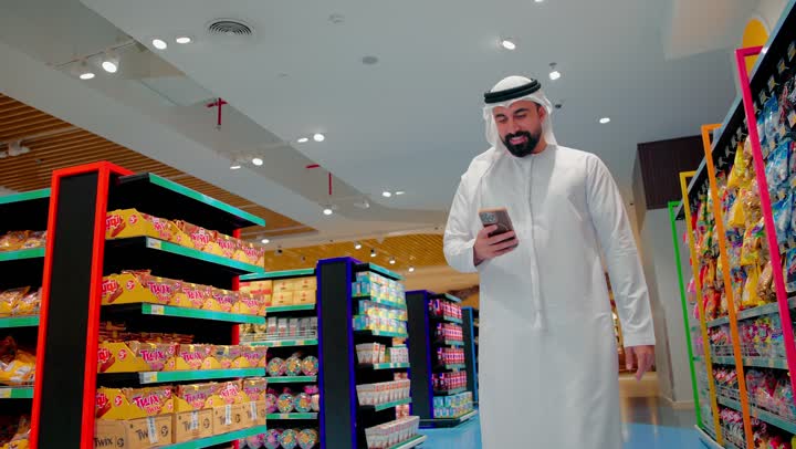 التواصل مع الاهل و الاصدقاء، تصفح مواقع التواصل الاجتماعي من خلال الجوال، رجل عربي خليجي اماراتي يرتدي الكندورة و الغترة يمشي داخل السوبر ماركت يقوم باستخدام الهاتف المحمول، مفهوم التسوق الذكي واستخدام التكنولوجيا في الحياة اليومية