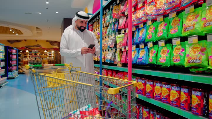 The concept of smart shopping and the use of modern technology in comparing products and making purchasing decisions, an Arab Gulf Emirati man wearing traditional attire stands in a supermarket aisle browsing his smartphone while shopping, shelves of canned food products, shopping inside modern stores.