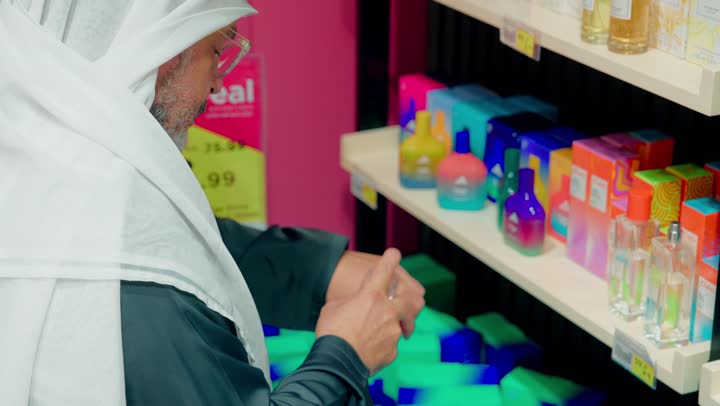 مستحضرات تجميل وعناية يومية، تصويرمقرب لرجل عربي خليجي اماراتي يرتدي الكندورة و الغترة يقف أمام رفوف مليئة بمنتجات العناية الشخصية والعطور داخل السوبر ماركت، فحص العبوات ومقارنة المنتجات قبل الشراء