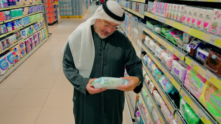 مقارنة المنتجات قبل الشراء، رجل عربي خليجي امارتي يرتدي الكندورة و الغترة داخل ممر السوبر ماركت يقوم بقراءة تفاصيل عبوة منتج، حرص المستهلك على اختيار المنتج المناسب، مفهوم التسوق الواعي