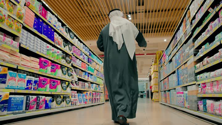 أرفف مليئة بالسلع الاستهلاكية والمنزلية ، اختيار المنتجات والبحث عن الجودة، تصوير من الخلف لرجل عربي خليجي اماراتي يرتدي الكندورة و الغترة يتجول داخل ممر السوبر ماركت يتفقد رفوف المنتجات المتنوعة، شراء الاحتياجات المنزلية