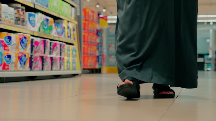 A close-up shot of an Emirati Gulf Arab man shopping in a supermarket aisle, acquiring daily essential supplies, moving between sections inside the store, with shelves full of various products.