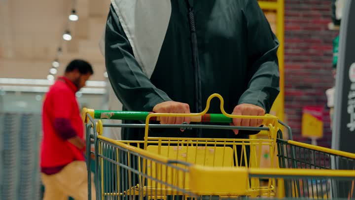A close-up shot of the hand of an Arab Gulf Emirati man holding a shopping cart inside a supermarket aisle, the concept of home shopping and selecting essential needs, shelves filled with various products, moving between sections inside the store.