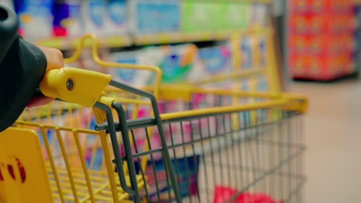 مفهوم التسوق المنزلي واختيار الاحتياجات الأساسية، تصوير مقرب ليد رجل عربي خليجي اماراتي يمسك عربة تسوق داخل ممر السوبر ماركت، رفوف مليئة بالمنتجات المتنوعة، التنقل بين الأقسام داخل المتجر