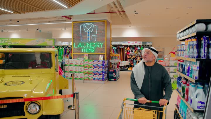 The concept of consumption and organizing household affairs, an Emirati Gulf Arab man wearing a kandura and ghutrah is strolling with a shopping cart in the laundry supplies section of the supermarket, selecting cleaning products and detergents from the shelves, comparing the types and offers available in the store, daily shopping, and purchasing household necessities.