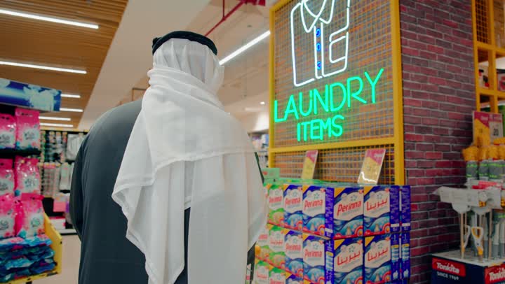 Buying household cleaning supplies, comparing products and offers inside the store, a rear view of an elderly Emirati Gulf Arab man choosing laundry powder and detergents from the supermarket shelves, daily shopping and attention to cleanliness, the concept of consumption and household needs.