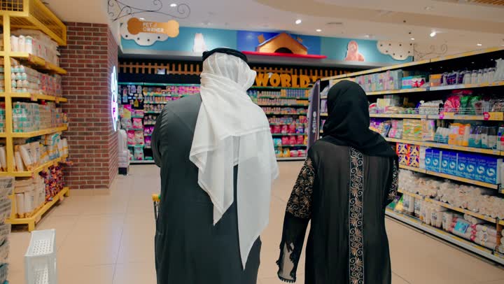 مفهوم التسوق العائلي والتخطيط للاحتياجات، شراء الاحتياجات المنزلية والمواد الاستهلاكية، تصوير من الخلف لرجل عربي خليجي اماراتي مسن يرتدي الكندورة و الغترة يقوم بالتسوق داخل سوبر ماركت برفقة ابنته، استخدام عربة التسوق داخل المتجر