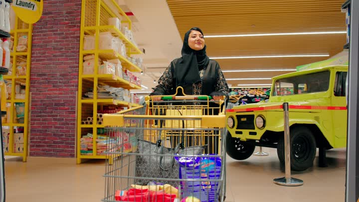 اختيار المواد الاستهلاكية والاحتياجات المنزلية، مفهوم الاستهلاك المنزلي والحياة اليومية، امرأة عربية خليجية اماراتية محجبة ترتدي العباءة تقوم بوضع منتج منظفات داخل عربة التسوق
