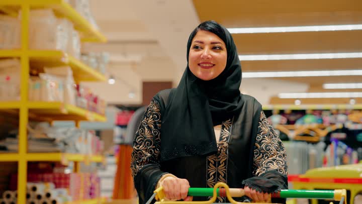 مفهوم الاستهلاك المنزلي والحياة اليومية، امرأة عربية خليجية اماراتية محجبة ترتدي العباءة تقوم بوضع منتج منظفات داخل عربة التسوق، اختيار المواد الاستهلاكية والاحتياجات المنزلية