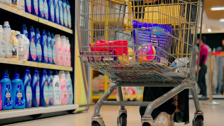 Preparing household supplies and needs, displaying food and household products on shelves, a close-up shot of an Emirati Gulf Arab woman pushing a shopping cart inside a supermarket aisle filled with products, the concept of consumer shopping.