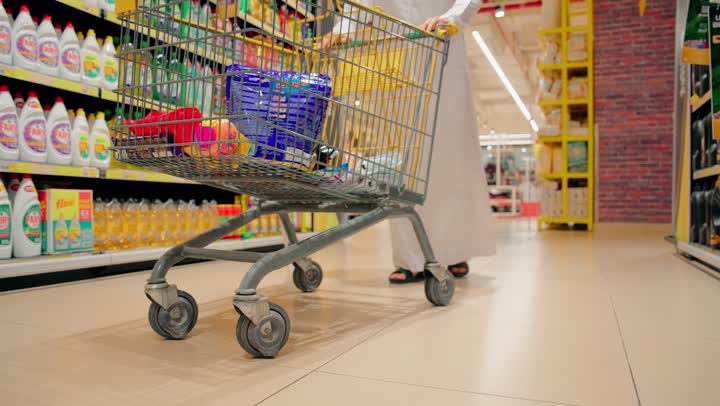 شراء الاحتياجات واللوازم المنزلية، تصوير مقرب لرجل عربي خليجي اماراتي يرتدي الزي التقليدي يجر عربة التسوق داخل السوبرماركت، الاستفادة من العروض والخصومات على المنتجات