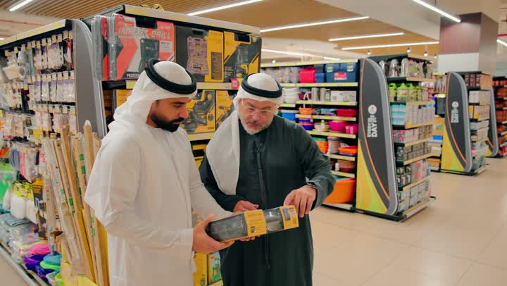 فحص عبوة منتج منزلي، تبادل الآراء أثناء التسوق داخل المركز التجاري، رجلان عربيان خليجيان اماراتيان يرتديان الكندورة و الغترة يتحدثان داخل متجر أدوات منزلية، مناقشة مواصفات منتج قبل الشراء، اختيار المستلزمات المنزلية