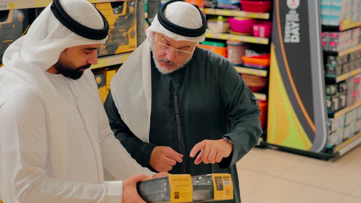 تبادل الآراء أثناء التسوق داخل المركز التجاري، تصوير مقرب لرجلان عربيان خليجيان اماراتيان يرتديان الكندورة و الغترة يتحدثان داخل متجر أدوات منزلية، مناقشة مواصفات منتج قبل الشراء، اختيار المستلزمات المنزلية، فحص عبوة منتج منزلي