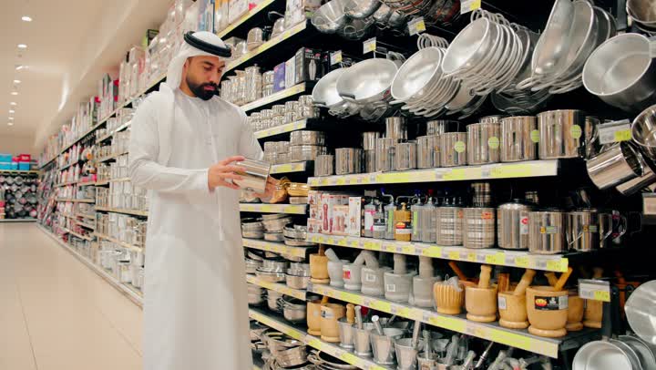 التأكد من جودة أدوات الطهي قبل الشراء، اختيار أواني مطبخ مصنوعة من الستانلس ستيل، رجل عربي خليجي اماراتي يرتدي الكندورة و الغترة يفحص وعاء معدني داخل متجر أدوات منزلية، التسوق داخل مركز تجاري، تجهيز المطبخ المنزلي بأدوات عملية
