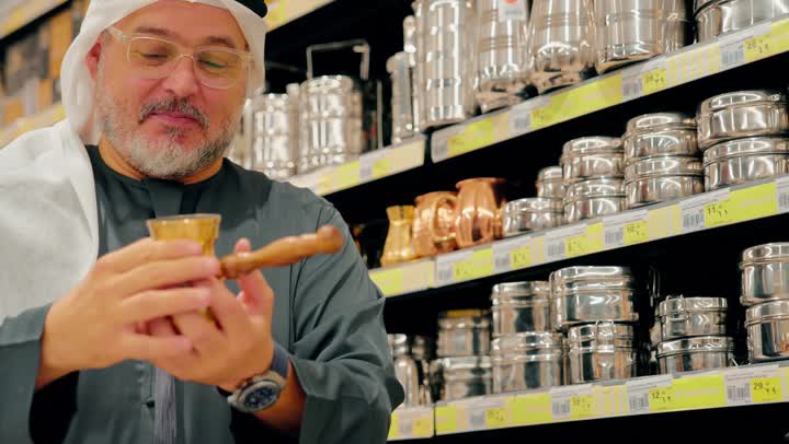 التسوق داخل المركز التجاري، فحص جودة الأدوات قبل الشراء، رجل عربي خليجي اماراتي  يرتدي الكندورة و الغترة يتسوق داخل متجر أدوات منزلية، تفقد أواني المطبخ المصنوعة من المعدن، اختيار مستلزمات الطهي للاستخدام اليومي، الاهتمام بتجهيز المطبخ المنزلي بأدوات عملية
