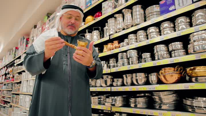 فحص جودة الأدوات قبل الشراء، رجل عربي خليجي اماراتي  يرتدي الكندورة و الغترة يتسوق داخل متجر أدوات منزلية، تفقد أواني المطبخ المصنوعة من المعدن، اختيار مستلزمات الطهي للاستخدام اليومي، التسوق داخل المركز التجاري، الاهتمام بتجهيز المطبخ المنزلي بأدوات عملية