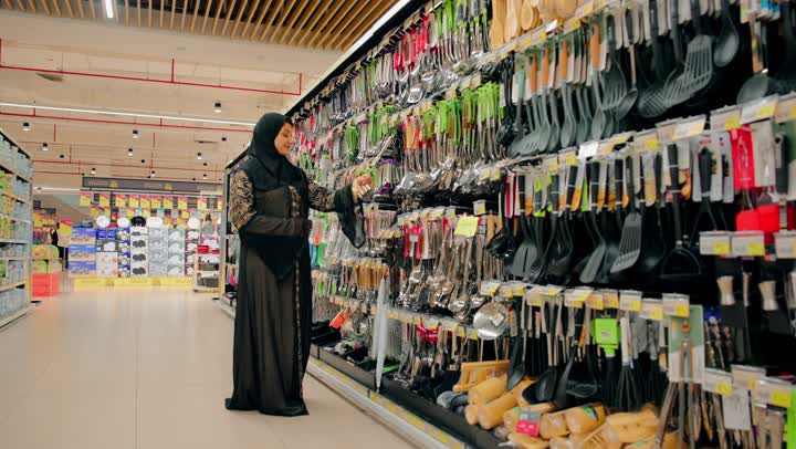 Choosing different kitchen tools, an Emirati Gulf Arab woman wearing an abaya and hijab is shopping inside a home goods store, checking out modern cooking supplies, shopping in the mall, focusing on preparing the home kitchen.