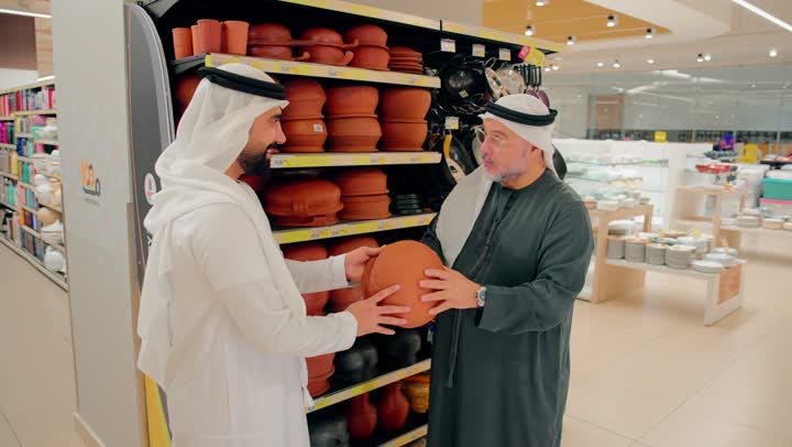 Exchanging opinions while choosing kitchen supplies, two Emirati Gulf Arab men dressed in traditional attire inspecting pottery inside a home goods store, interest in heritage tools made of pottery, shopping inside a shopping mall.