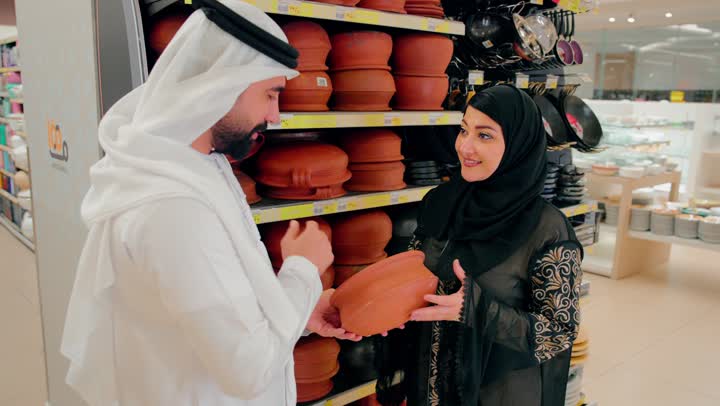 التعاون في اختيار مستلزمات المطبخ، الاهتمام بالطهي الصحي والأدوات التراثية، زوجان عربيان خليجيان اماراتيان يرتديان الزي التقليدي يتفقدان أواني فخارية تقليدية داخل متجر أدوات منزلية، اختيار أواني طبخ مصنوعة من الفخار الطبيعي، التسوق المشترك داخل المركز التجاري