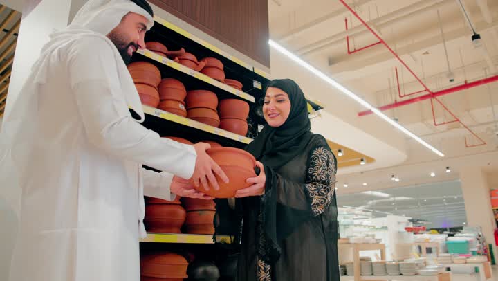 الاهتمام بالطهي الصحي والأدوات التراثية، زوجان عربيان خليجيان اماراتيان يرتديان الزي التقليدي يتفقدان أواني فخارية تقليدية داخل متجر أدوات منزلية، اختيار أواني طبخ مصنوعة من الفخار الطبيعي، التسوق المشترك داخل المركز التجاري، التعاون في اختيار مستلزمات المطبخ