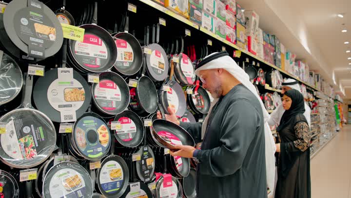 The focus on equipping the kitchen with practical tools, participating in the selection of kitchen utensils, family cooperation and bonding, an Emirati Gulf Arab family dressed in traditional attire choosing cooking pots inside a home goods store, the concept of family shopping within a commercial center.